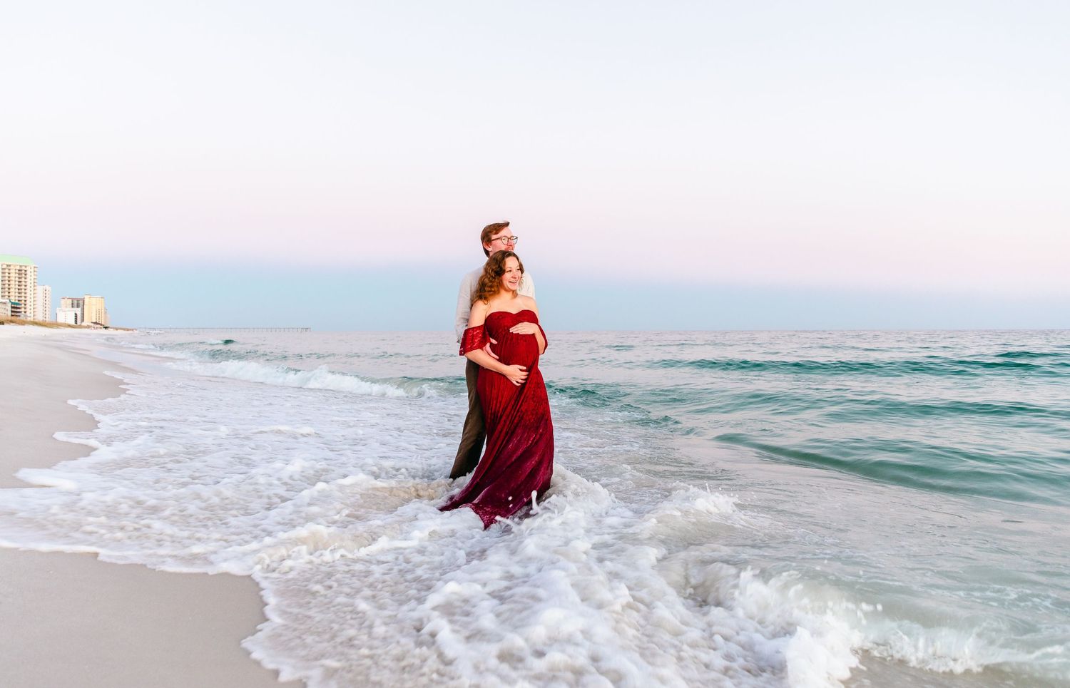 Elegant maternity photo of woman in flowing red dress standing in ocean waves at sunset.