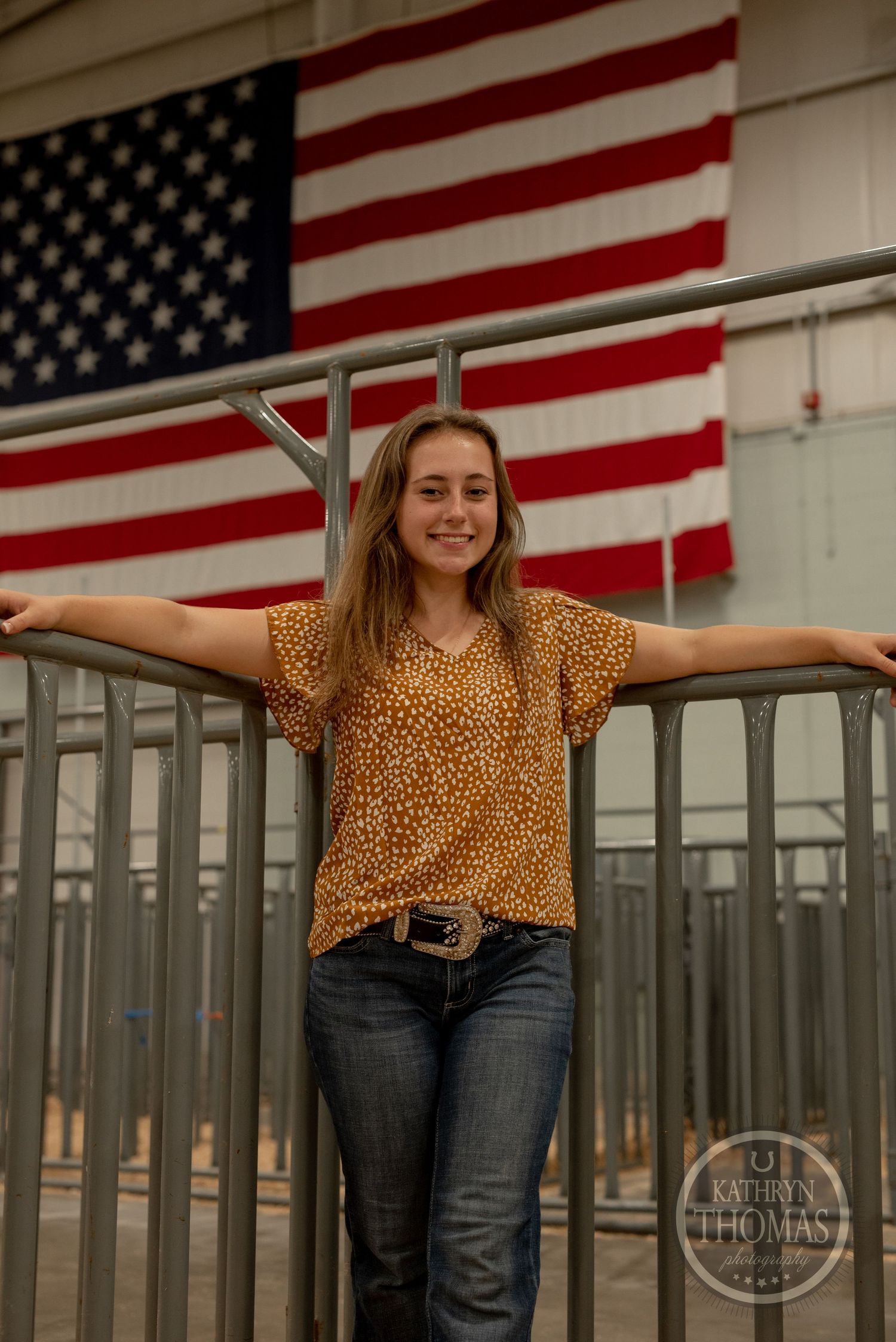 Casual pose against American flag backdrop in a livestock facility wearing spotted shirt and jeans.