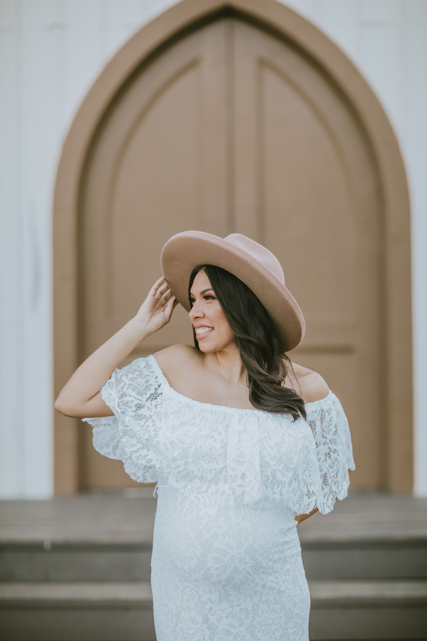 A radiant maternity portrait featuring a white lace dress and tan hat against an arched doorway.