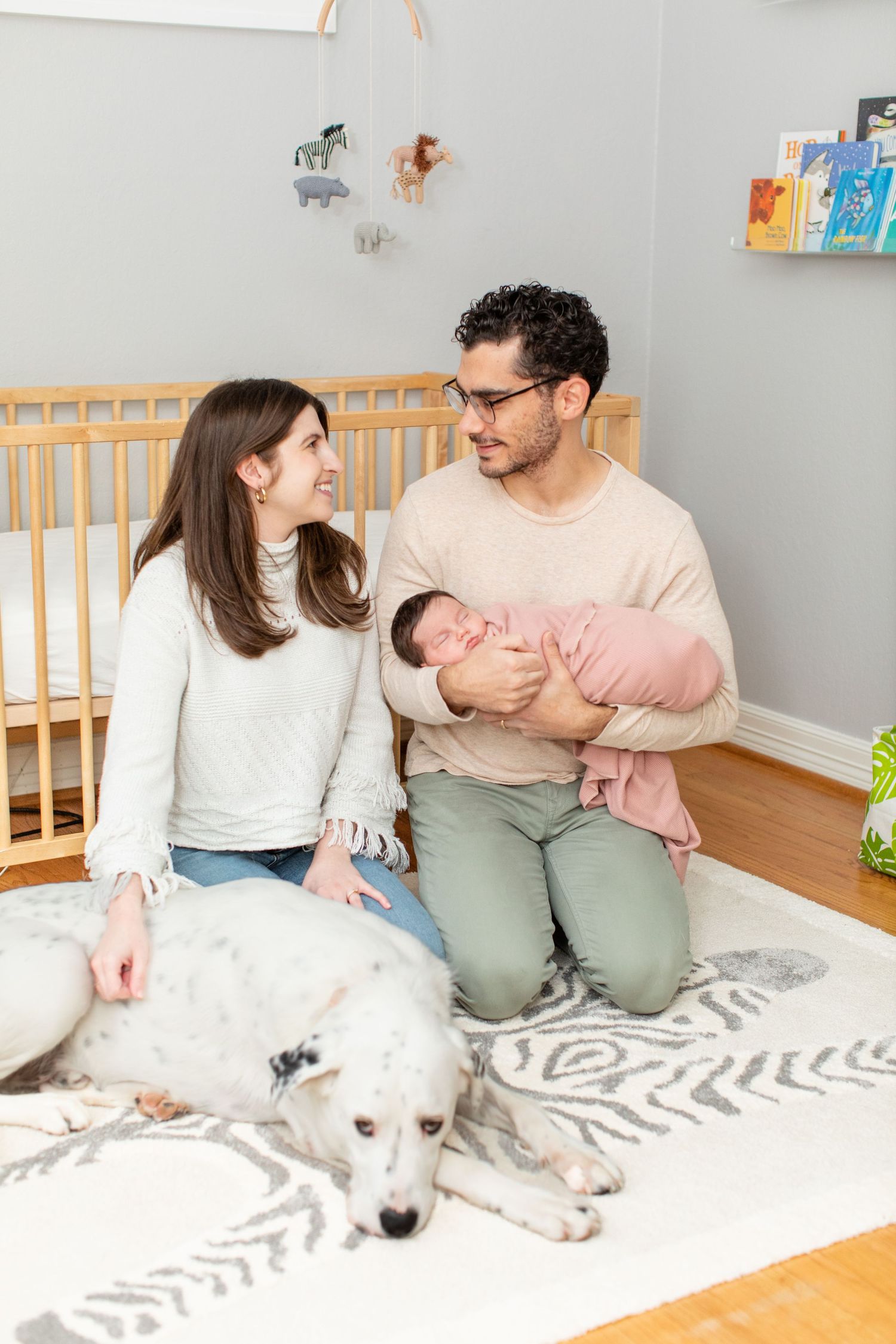 Family moment in nursery with white dog as parents look at each other on zebra print rug.