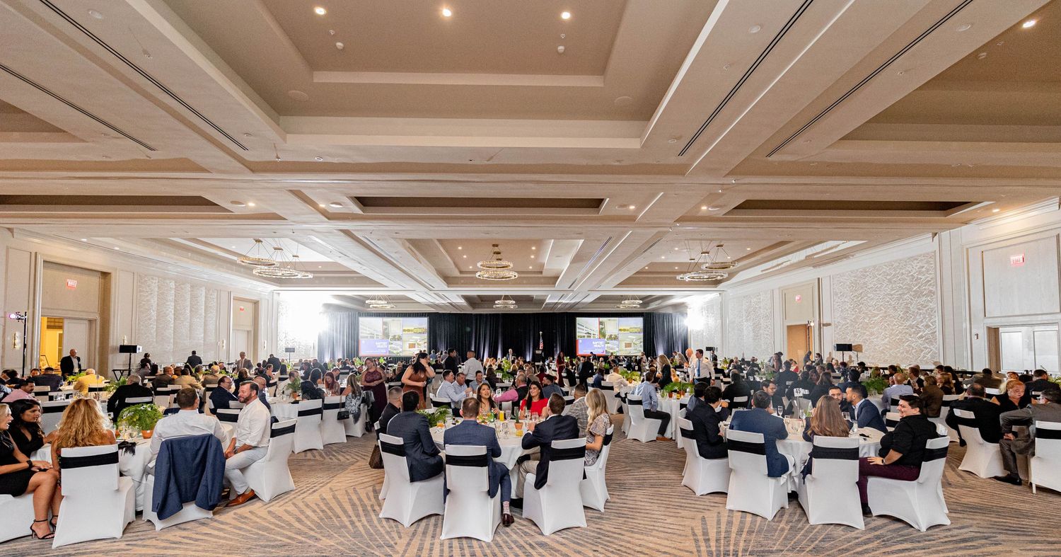 Elegant ballroom event with round white-draped tables and guests seated facing a large presentation screen.
