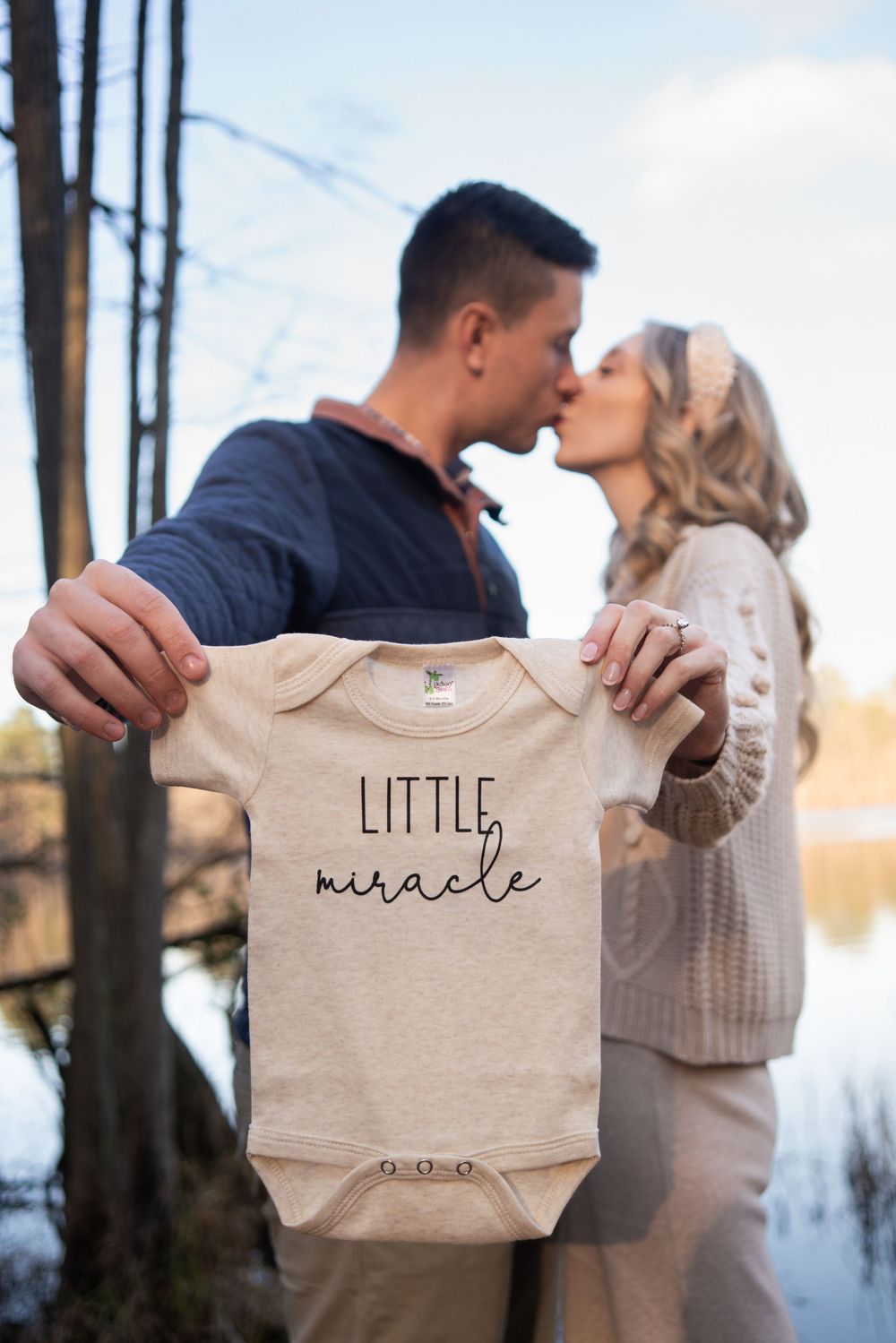 A couple holds up a baby onesie with 'Little Miracle' text during a pregnancy announcement photoshoot.