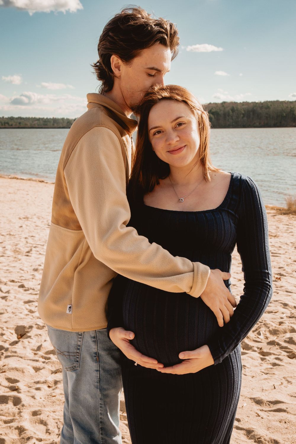 A romantic maternity photo of a couple embracing on a sandy beach with a lake and forest in the background.