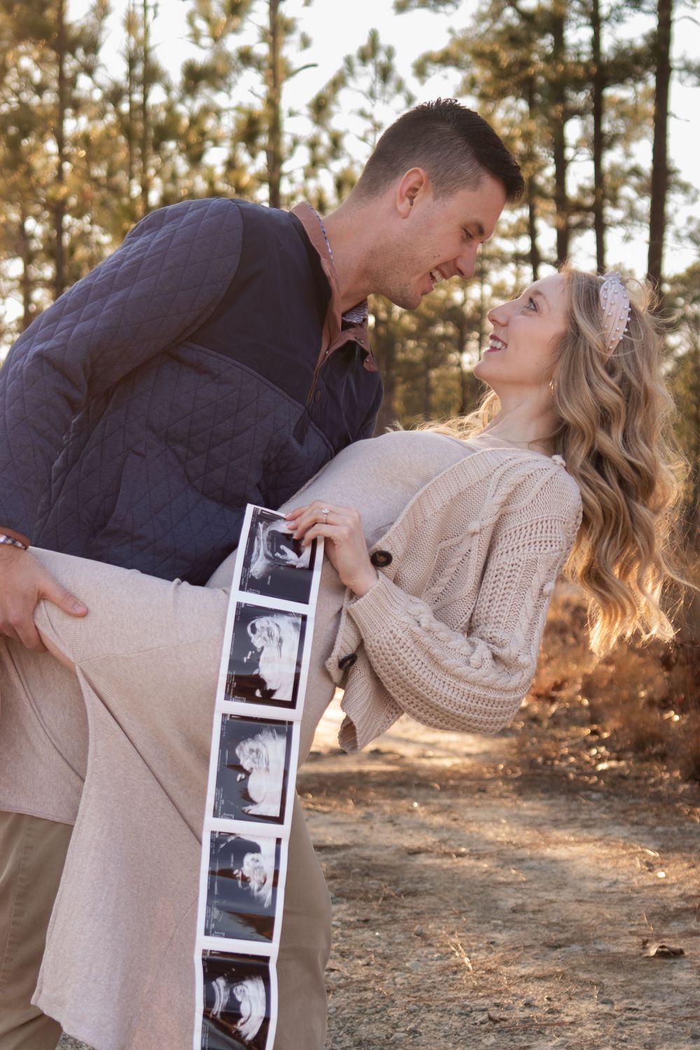 A couple shares an intimate moment while holding ultrasound photos in a wooded setting.