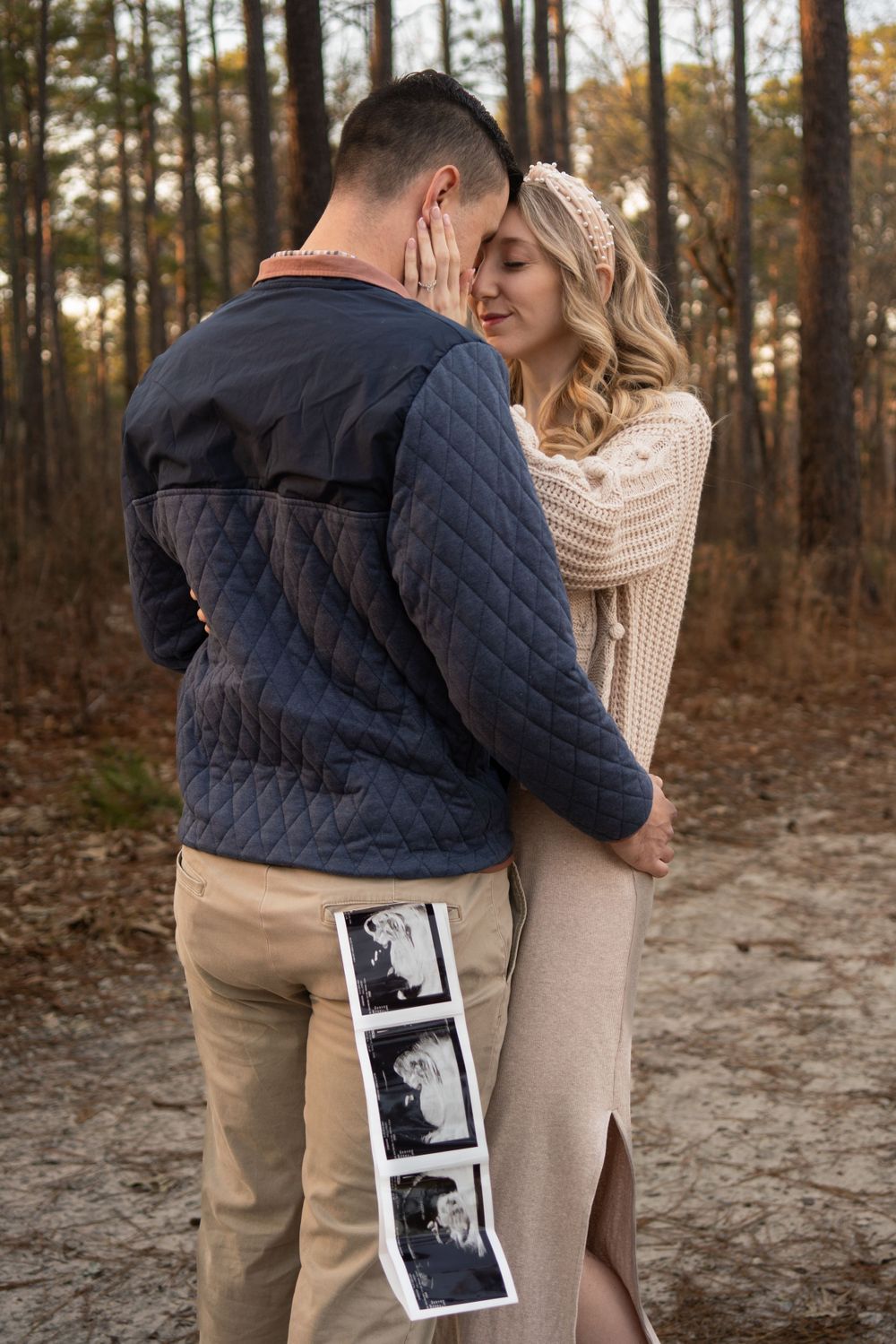 A couple embraces while holding ultrasound photos in a forest during sunset.
