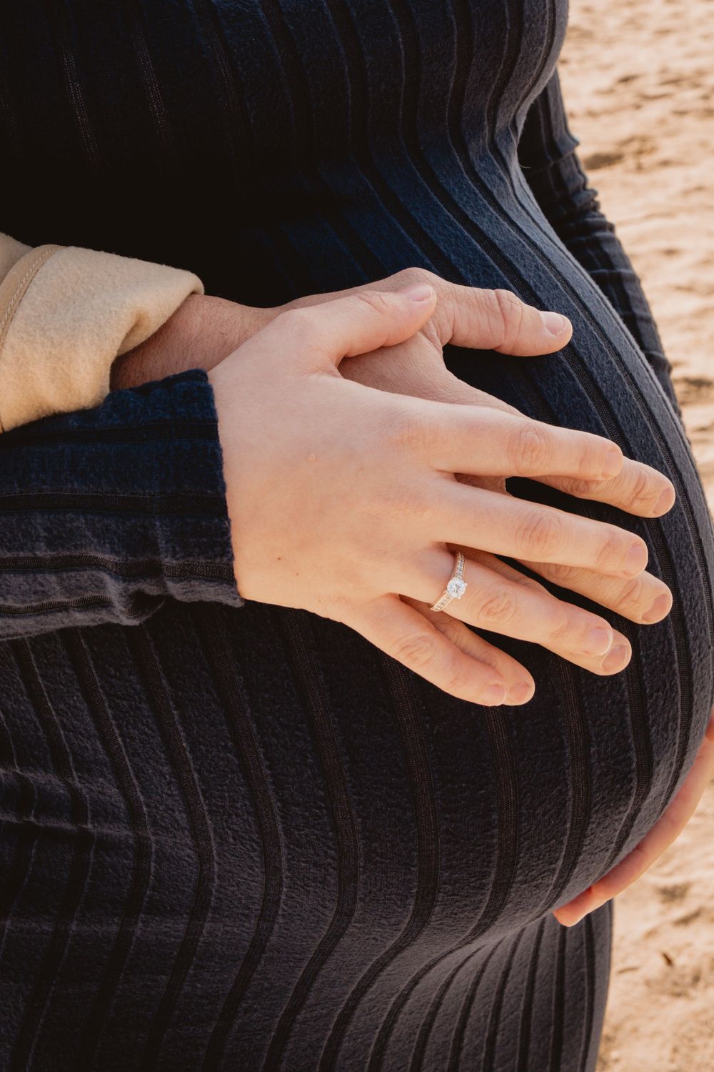 Close-up of hands tenderly cradling a pregnant belly in a black ribbed dress on a beach.