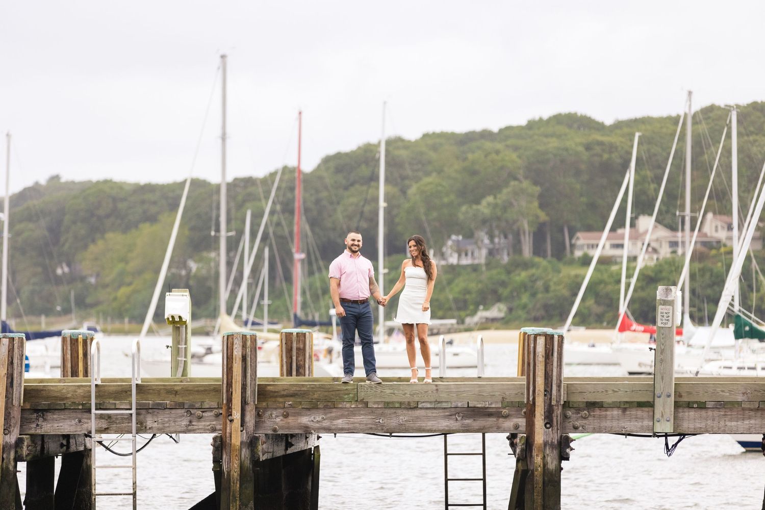 A couple stands together on a wooden dock overlooking a marina with boats.