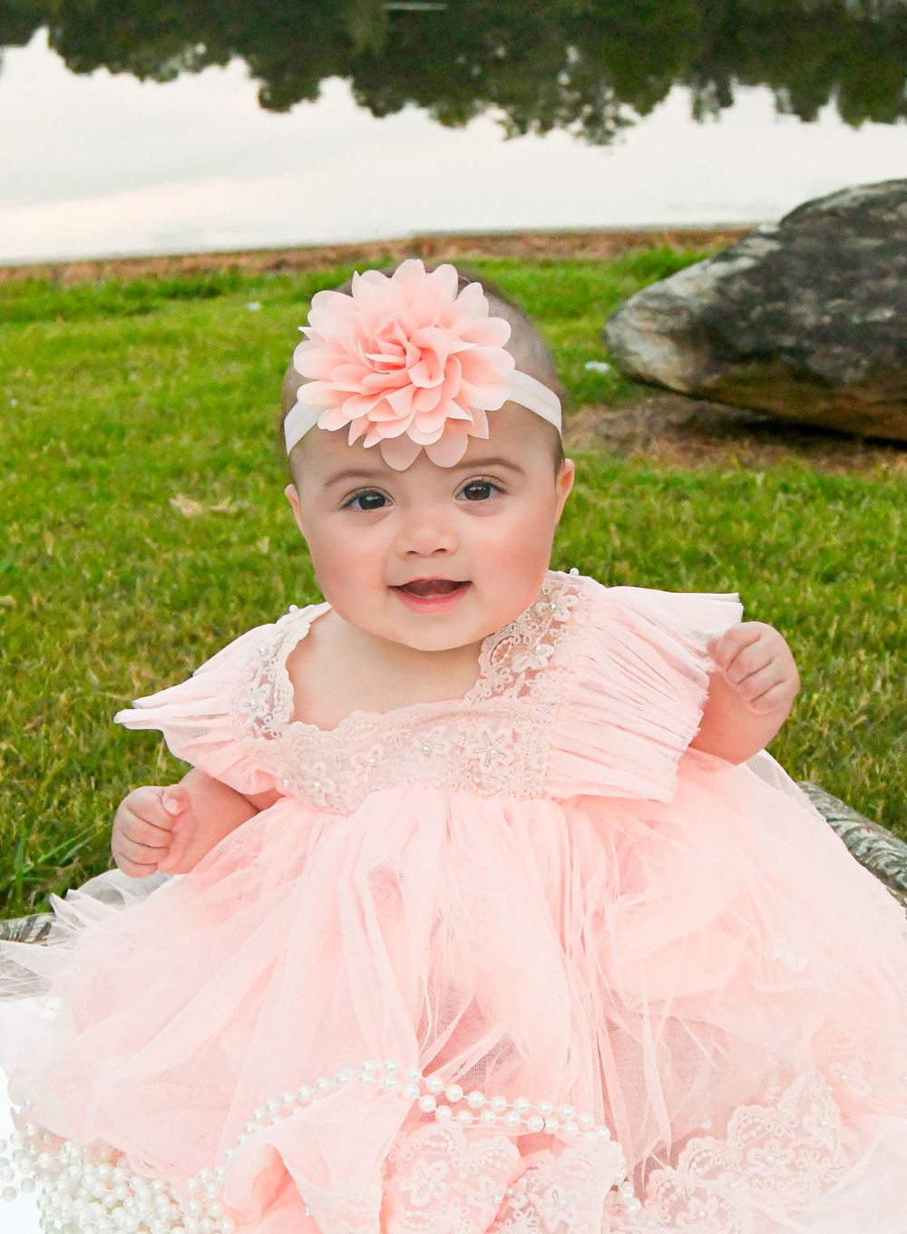 Baby in a frilly peach dress and headband sitting on grass.