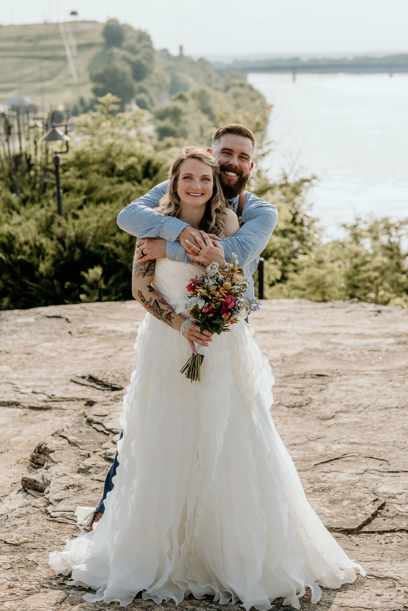 A newlywed couple embraces on a cliff overlooking a scenic lake view.