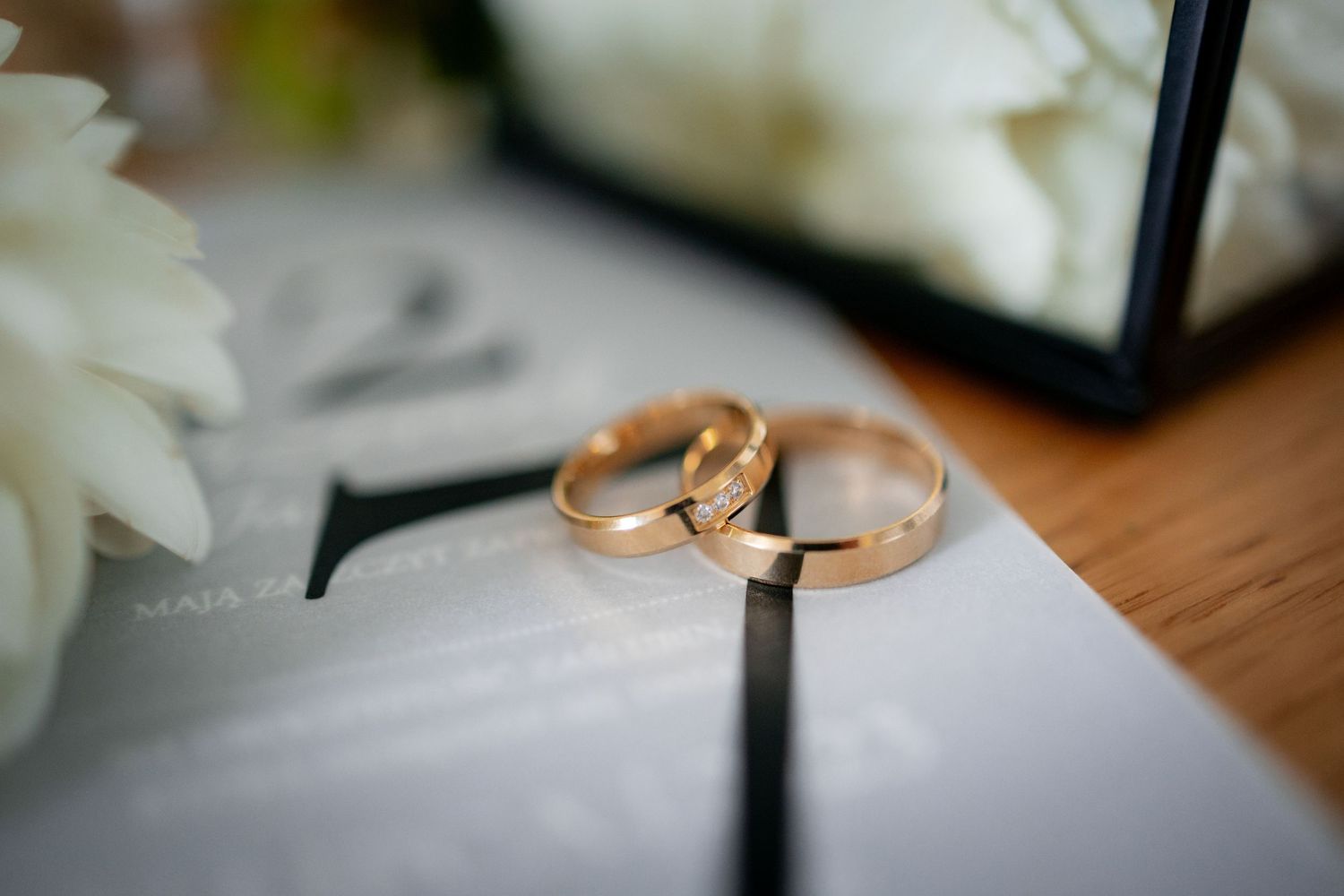 Two golden wedding rings placed on white paper with soft focus flower in background.