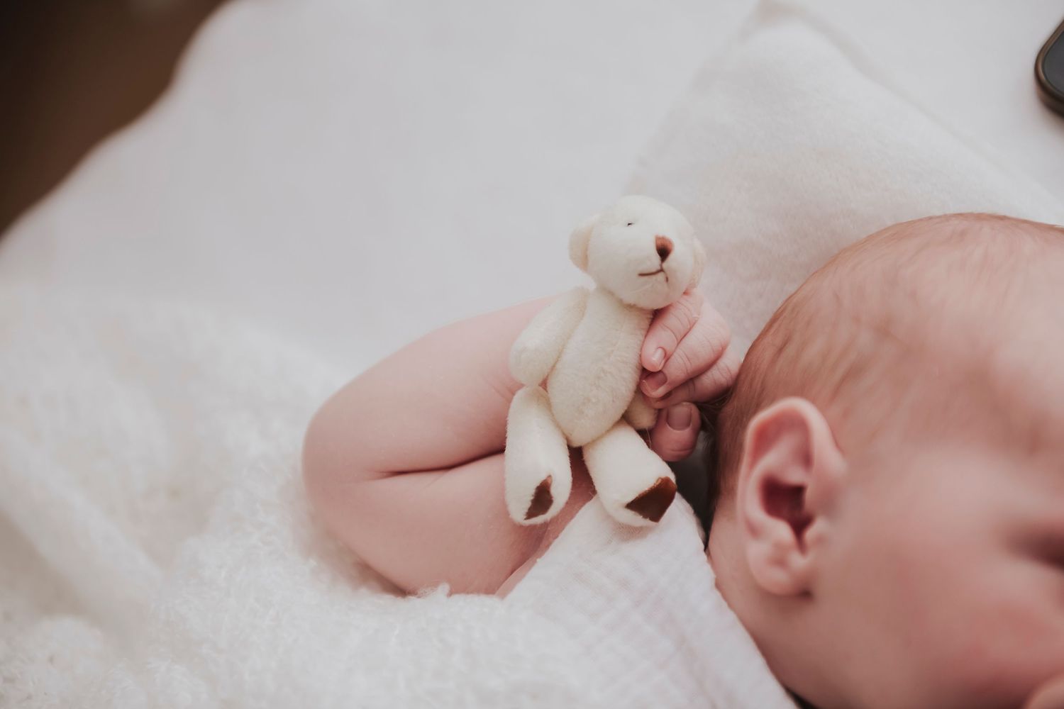 A sleeping infant cuddles with a small white teddy bear on white bedding.