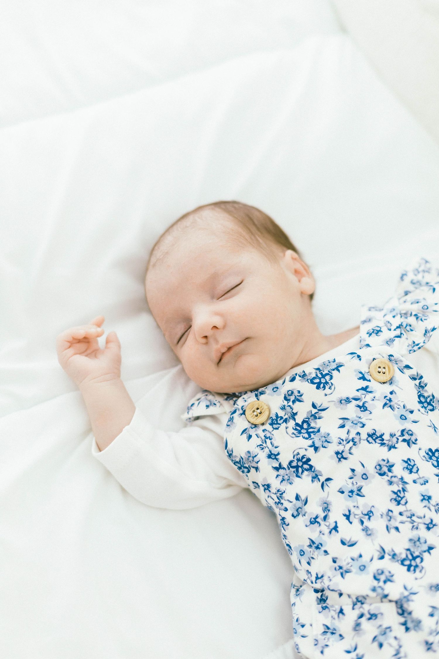 Infant wearing white and blue floral sleepwear naps on soft white bedding.