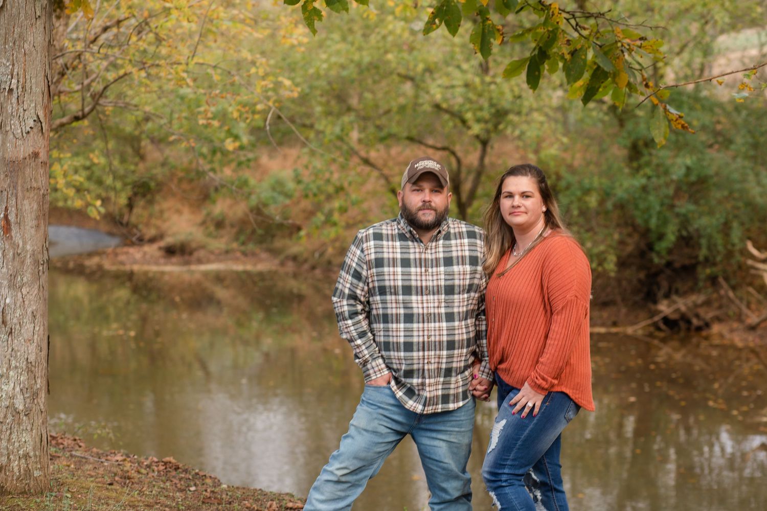 Couple sits together on wooden bench surrounded by autumn foliage near a peaceful pond.