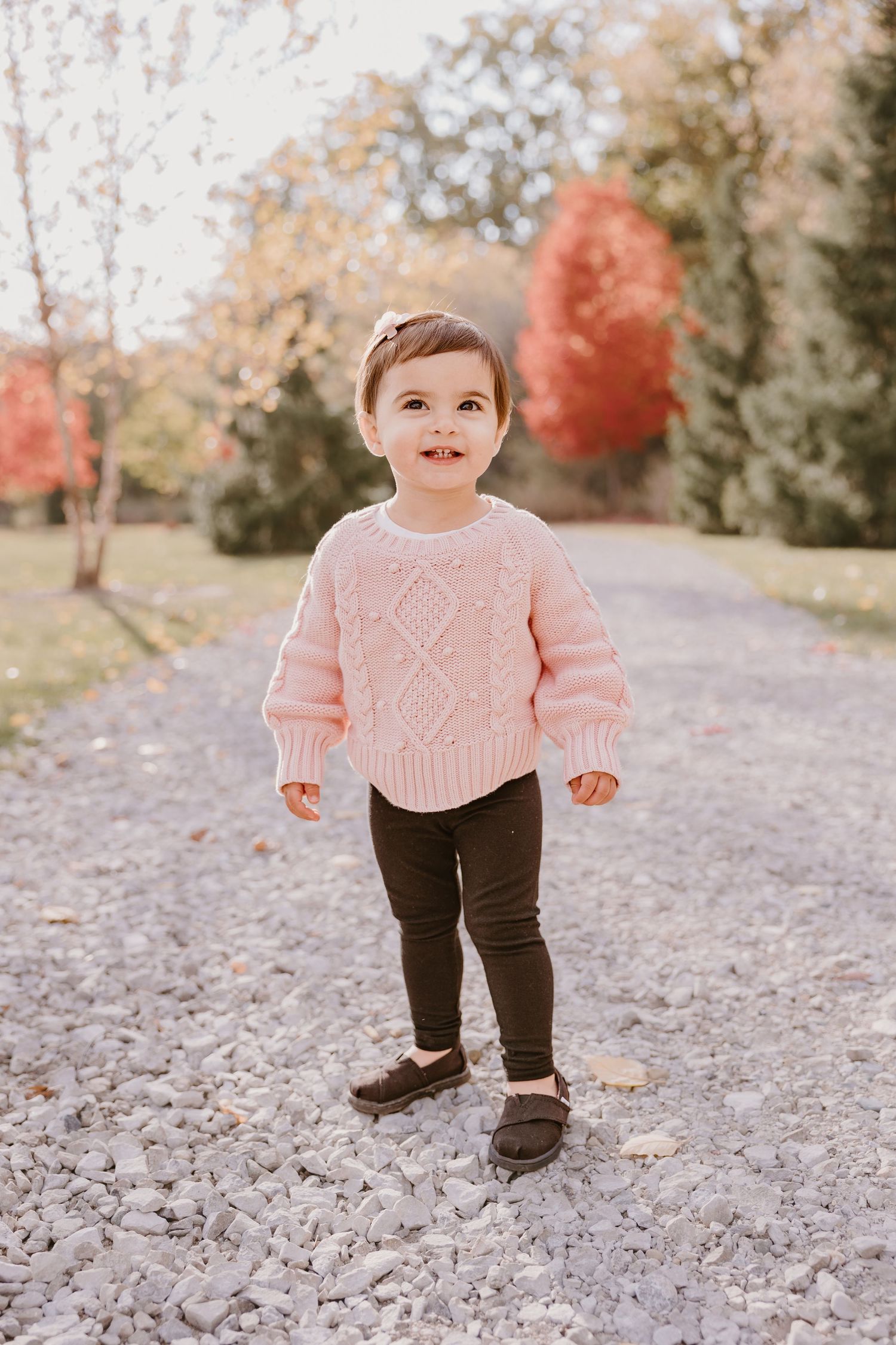 A small child wearing a pink sweater and dark pants stands on a gravel path in an autumn setting.
