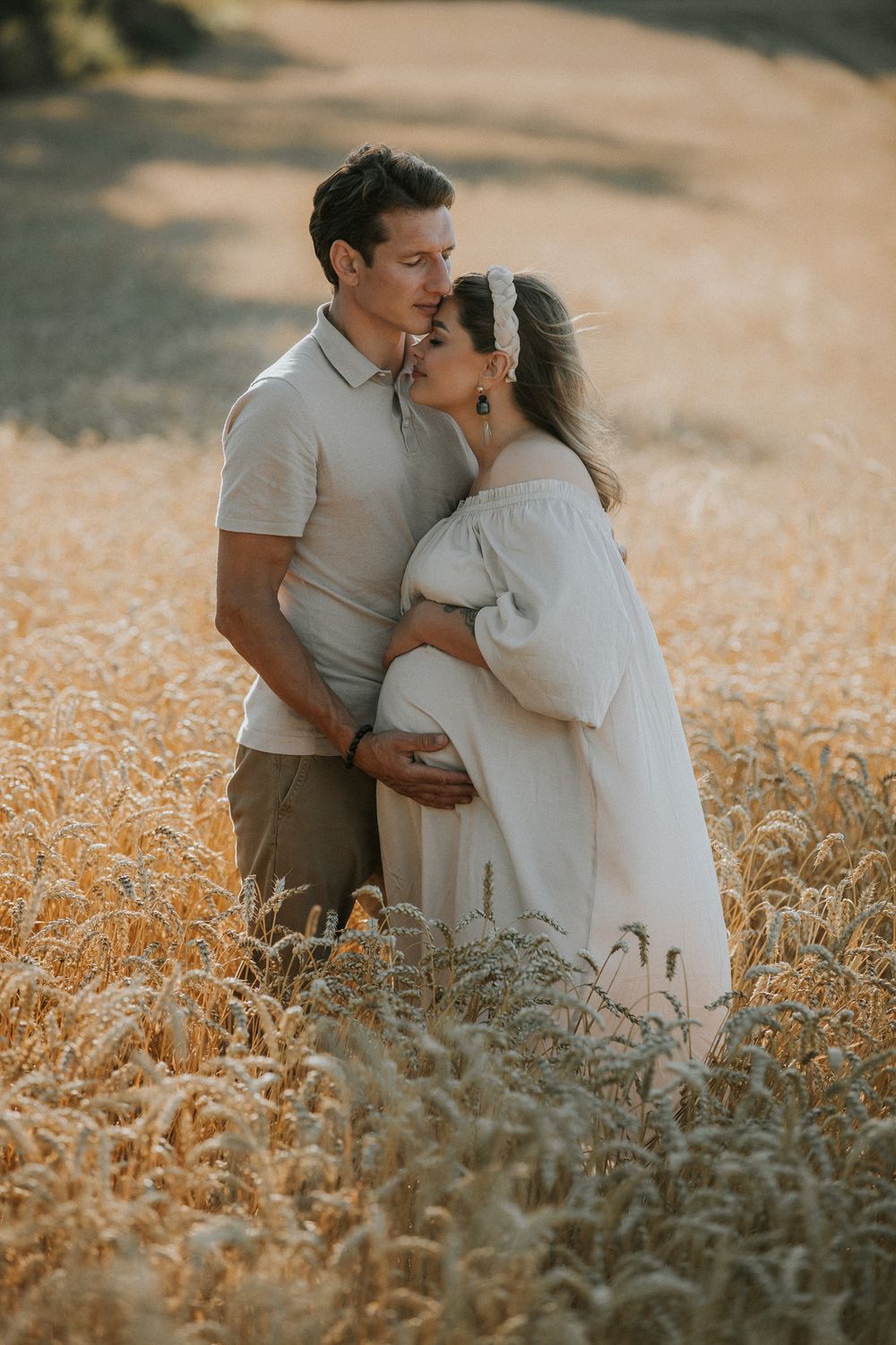 Couple sharing a tender moment in a golden wheat field, man kissing pregnant woman's forehead during maternity photoshoot.