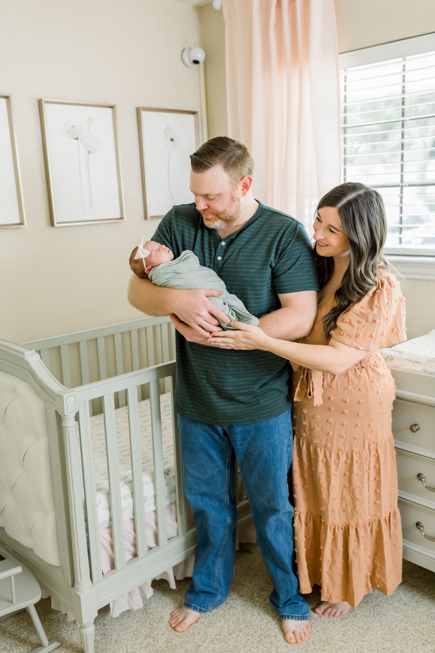 A couple holding their newborn baby next to a gray crib in a softly lit nursery.