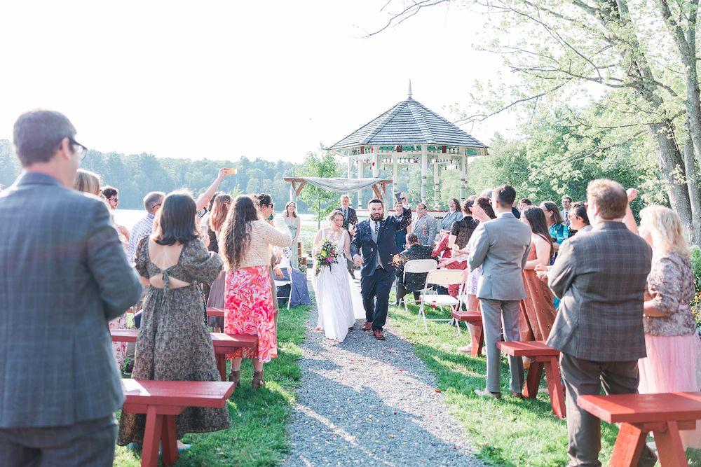 Outdoor wedding ceremony setup with red benches and guests gathering near a rustic gazebo.