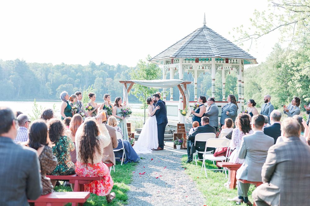 Scenic lakeside wedding ceremony with guests seated on wooden chairs around octagonal gazebo.