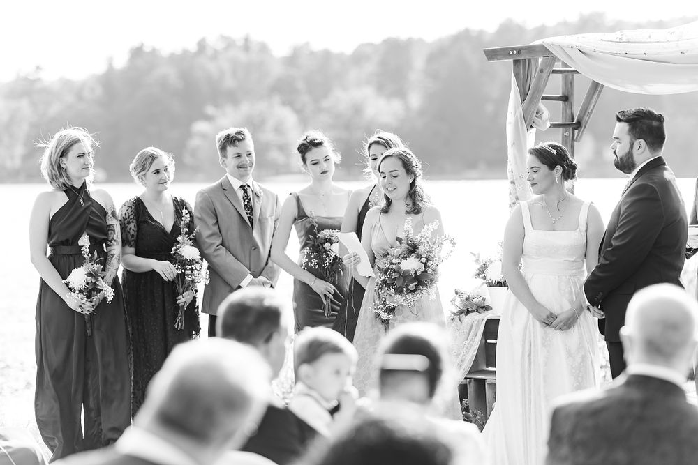 Black and white photo of wedding party standing at waterfront ceremony location.