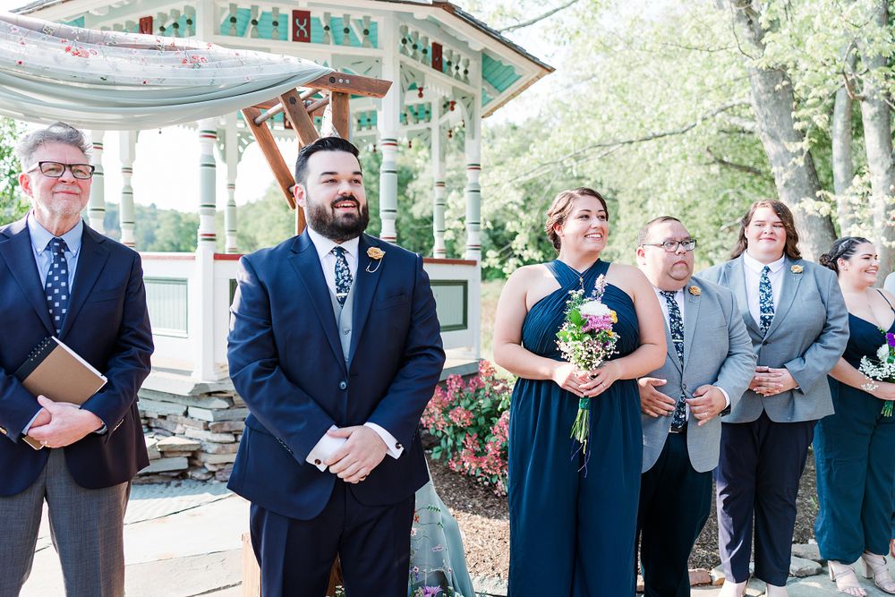 Wedding party stands under a gazebo during an outdoor ceremony with attendants in navy blue and teal attire.