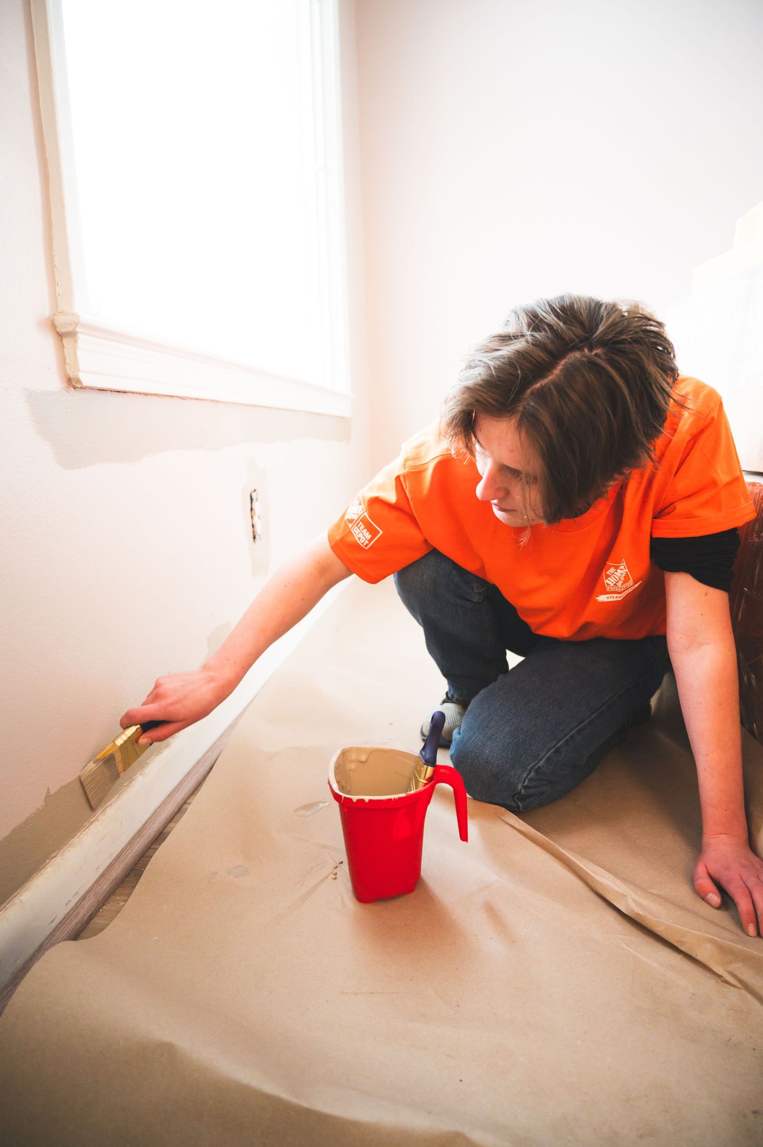 Person in orange t-shirt working on baseboard trim with paint supplies.