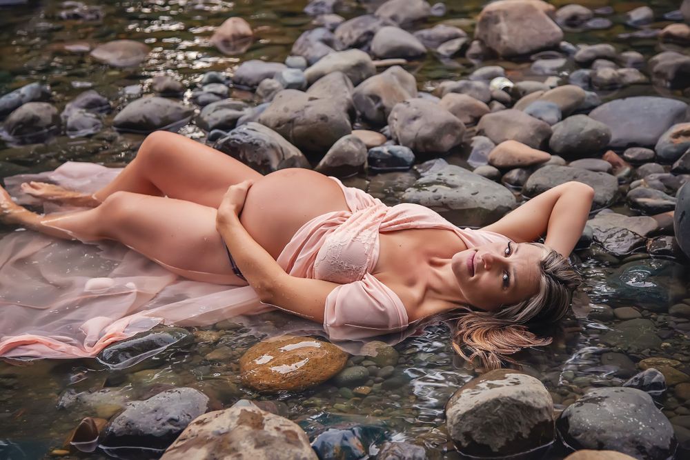 A pregnant woman lies in the bed of Puntledge River among the rocks wearing a pink dress
