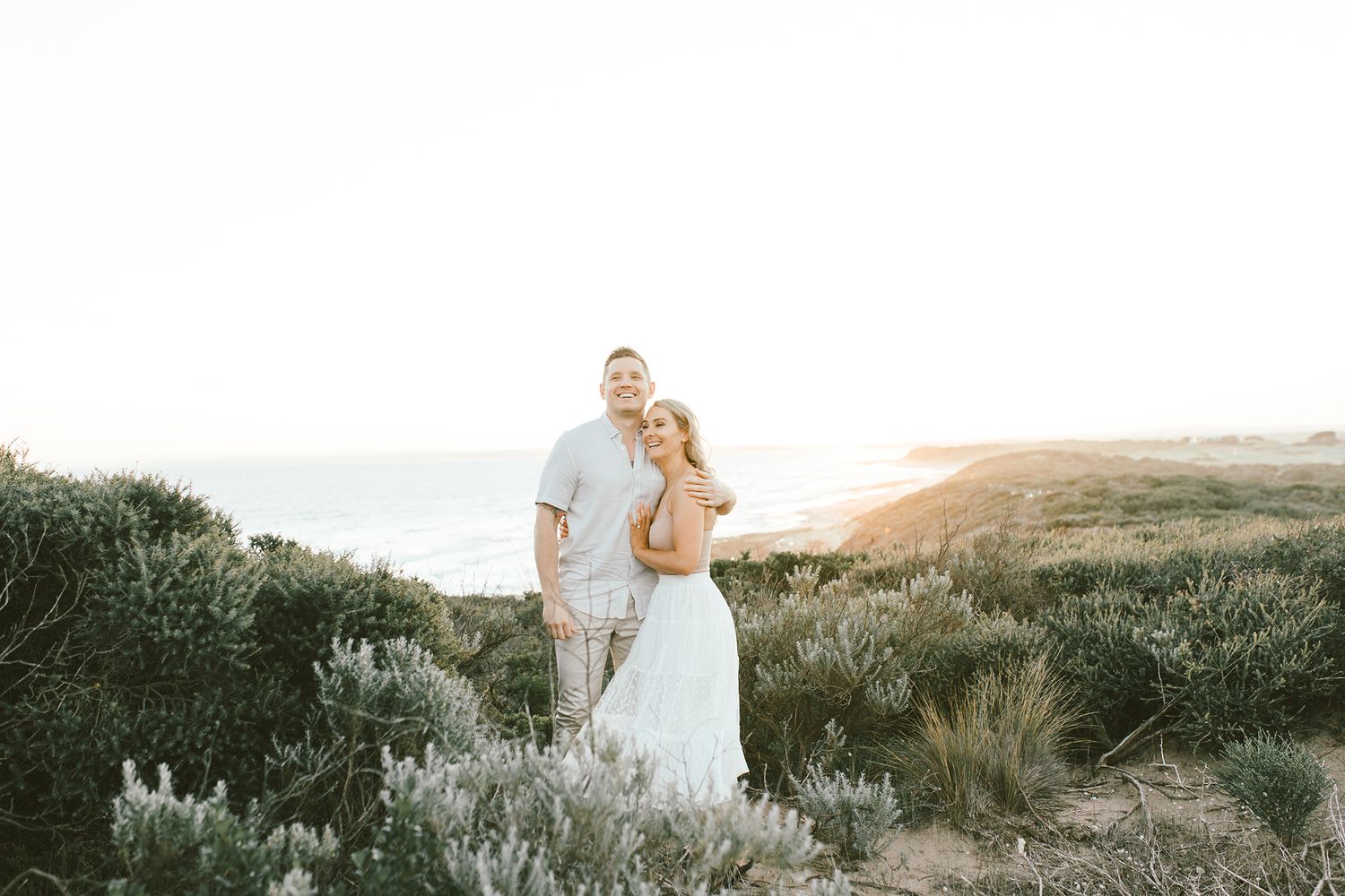 Romantic couple embracing on coastal cliffs during a dreamy golden hour photoshoot.