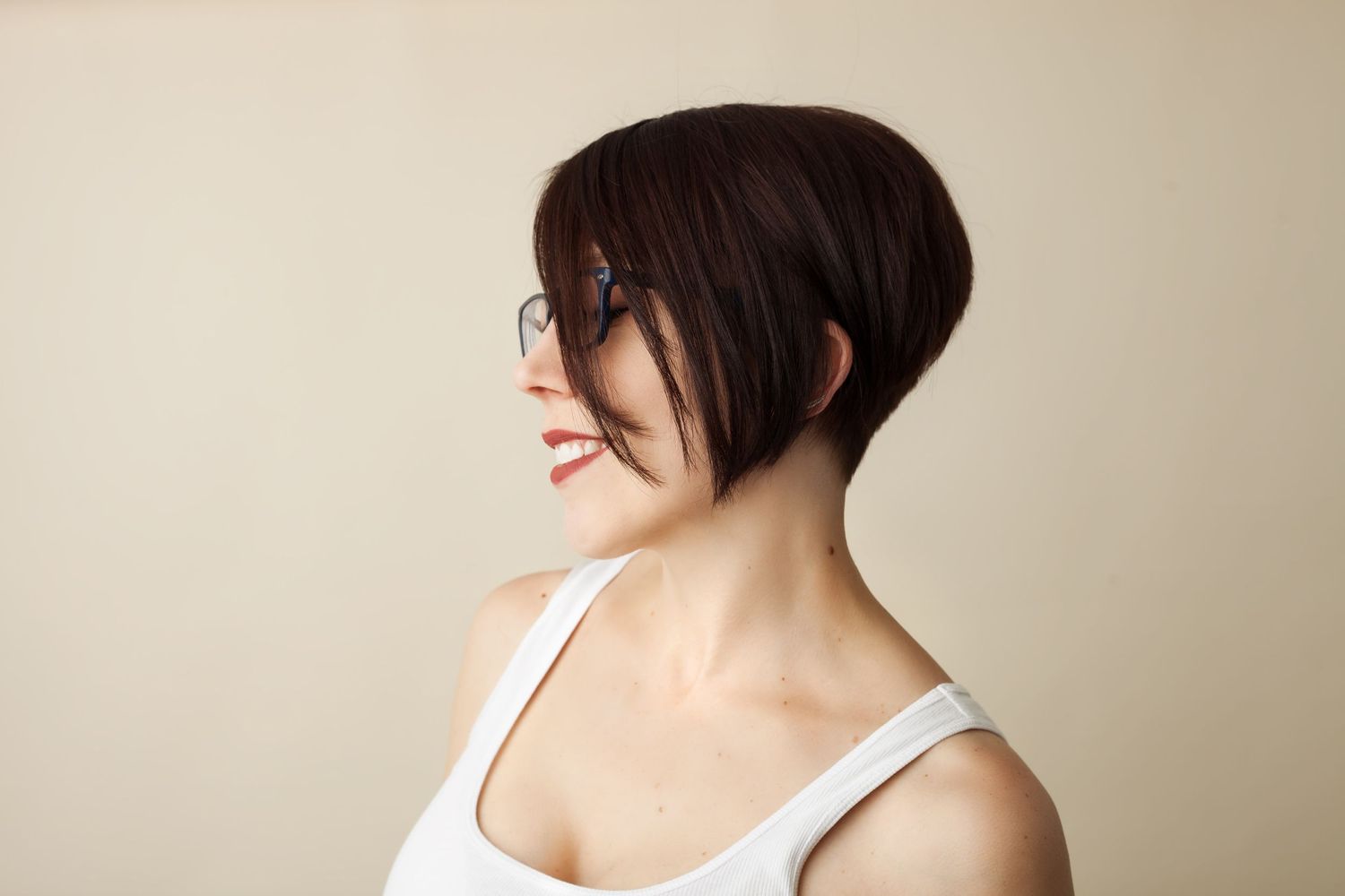 Side profile of a person with short dark hair wearing a white tank top smiling against beige background.