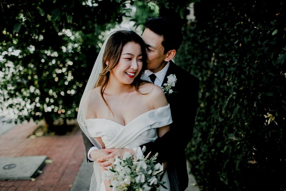 groom kissing bride on cheek next to grass wall