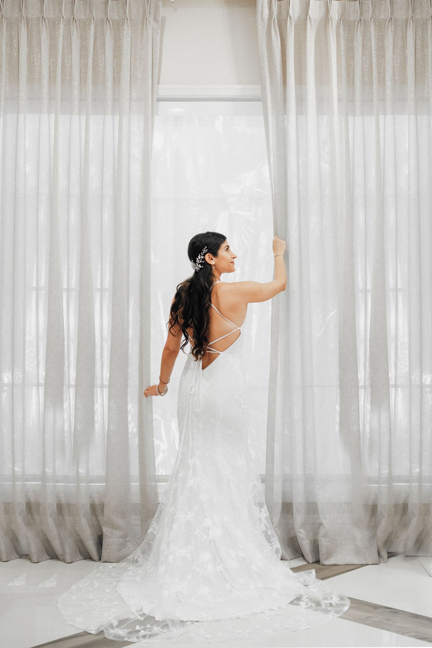 Bride in form-fitting lace gown poses elegantly against white curtains.