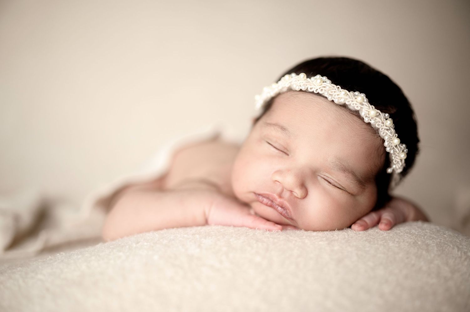 Peaceful sleeping newborn wearing delicate white headband on soft beige backdrop.