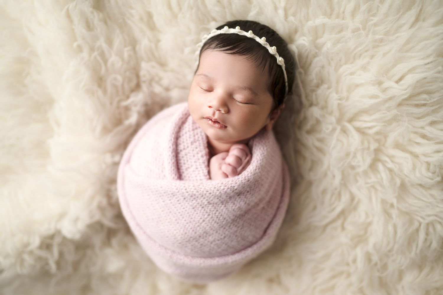 Sleeping baby swaddled in pink wrap on soft white fur background.