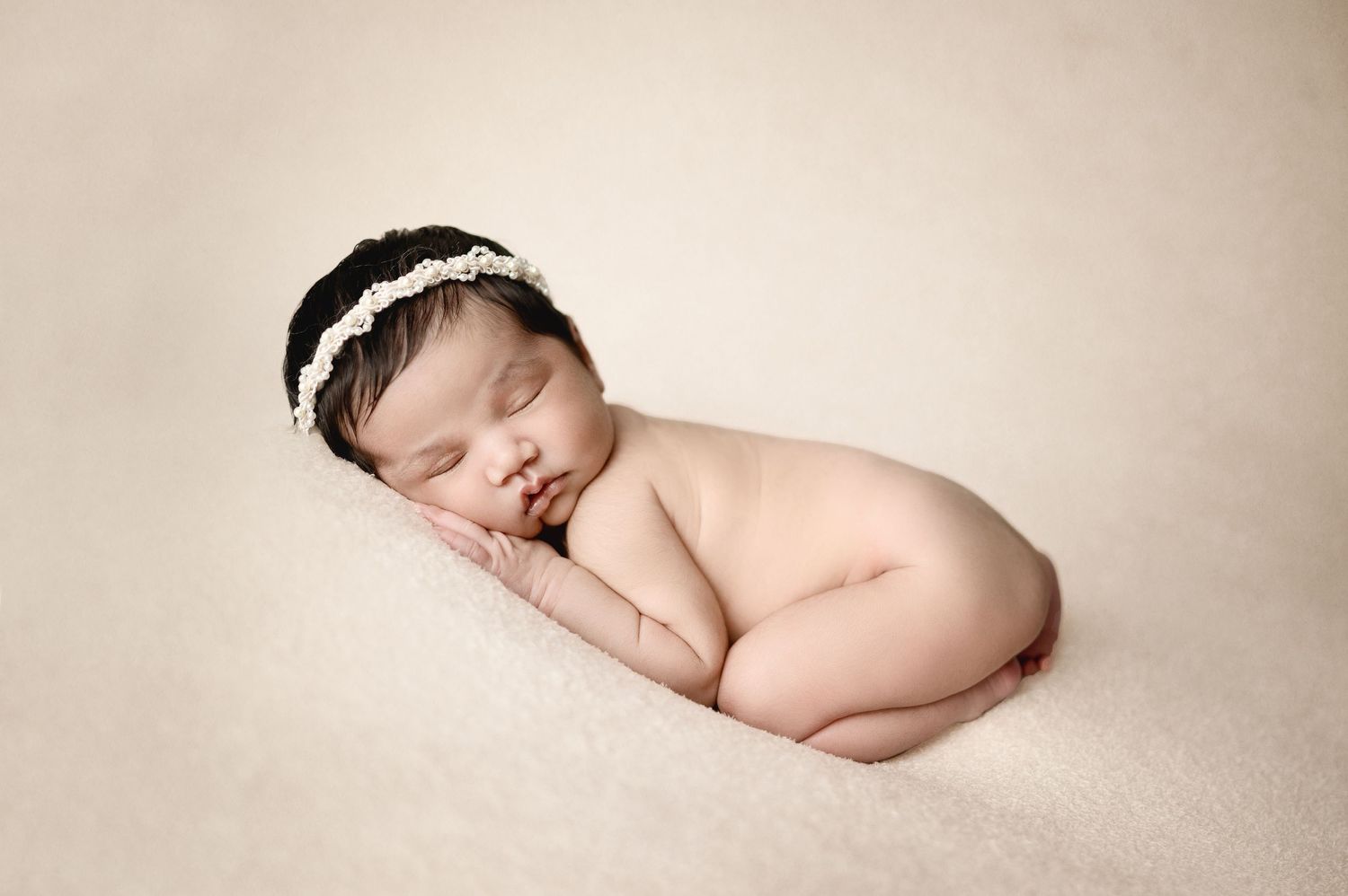 Newborn baby sleeping in fetal position with white lace headband on beige backdrop.