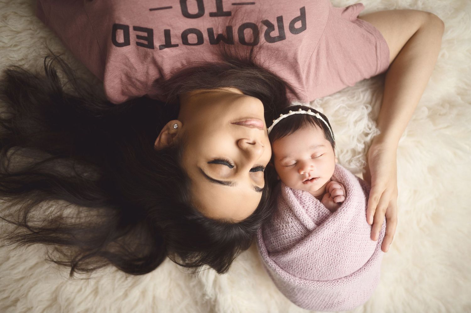 Mother and newborn portrait on white fur with baby wrapped in pink swaddle.