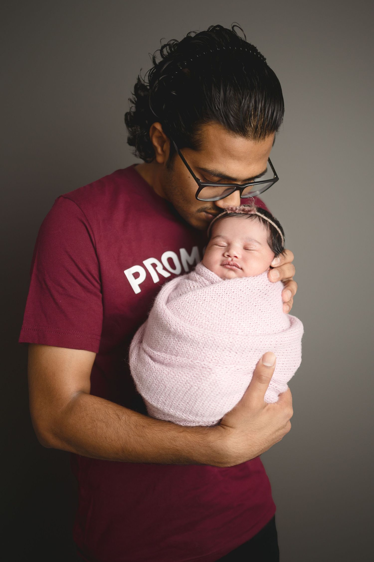 Parent in burgundy shirt holding newborn baby swaddled in pink blanket against grey background.