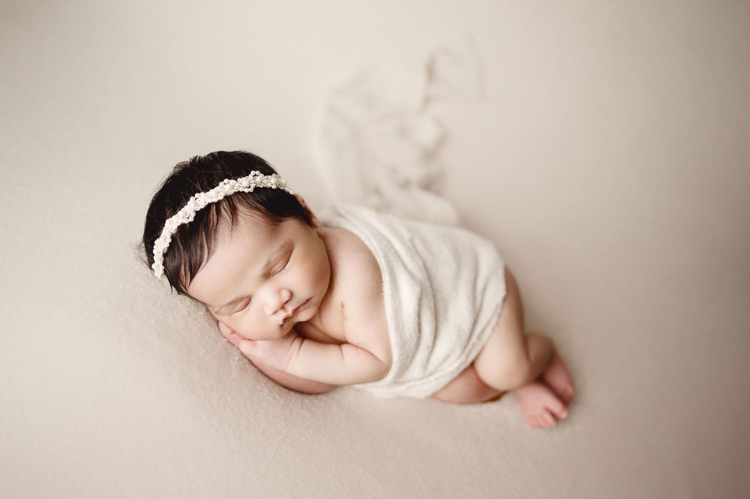 Peaceful newborn sleeping while wrapped in soft white fabric on neutral background.