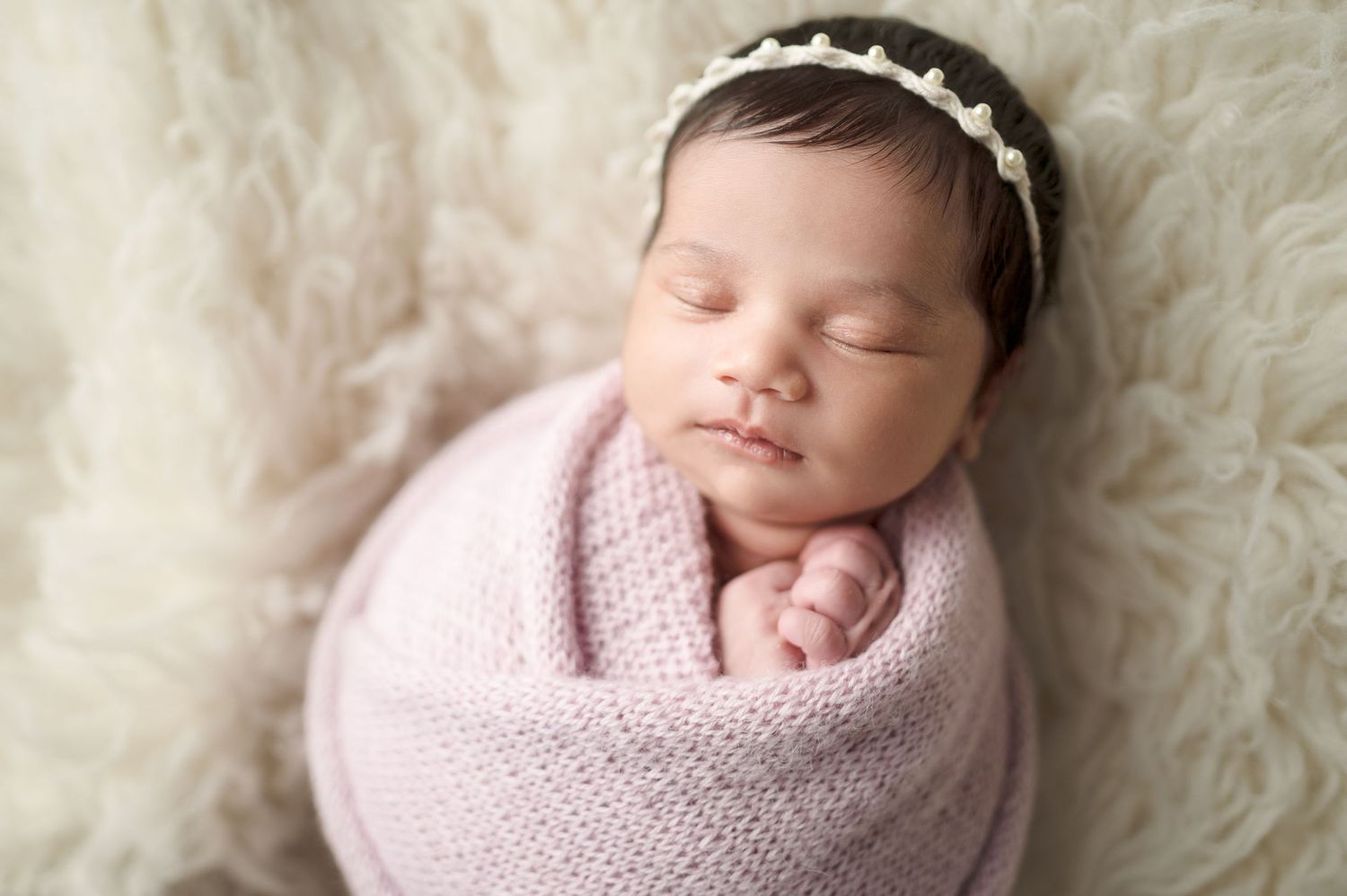 Newborn wrapped in pink knit blanket sleeping on fluffy white fur.