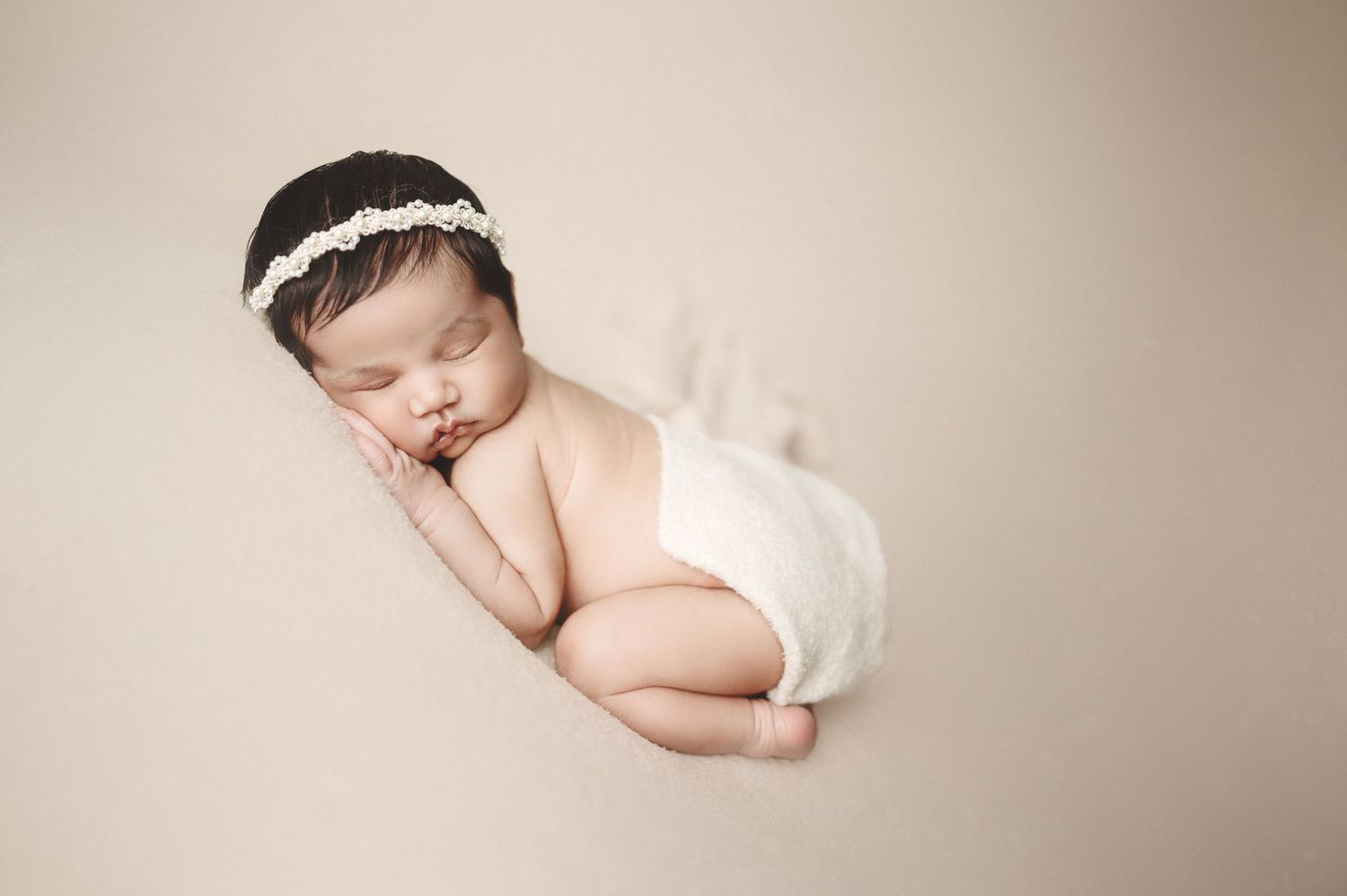 Sleeping newborn wearing white headband and diaper on minimalist beige backdrop.