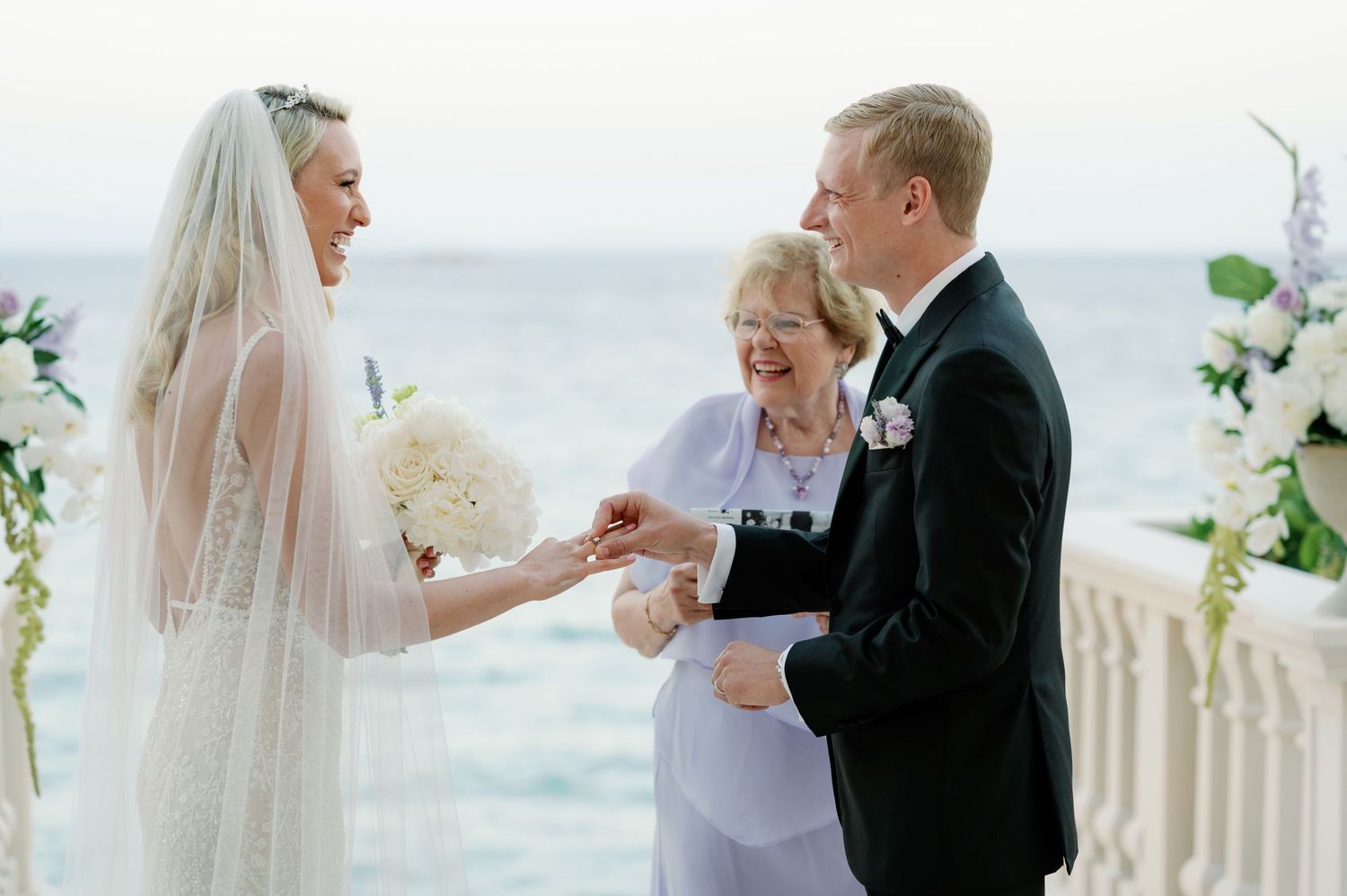 Ring exchange during seaside wedding ceremony with elegant white floral decor.