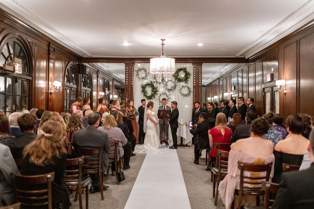 Indoor wedding ceremony with wooden chairs, chandeliers, and holiday wreaths adorning the paneled walls.