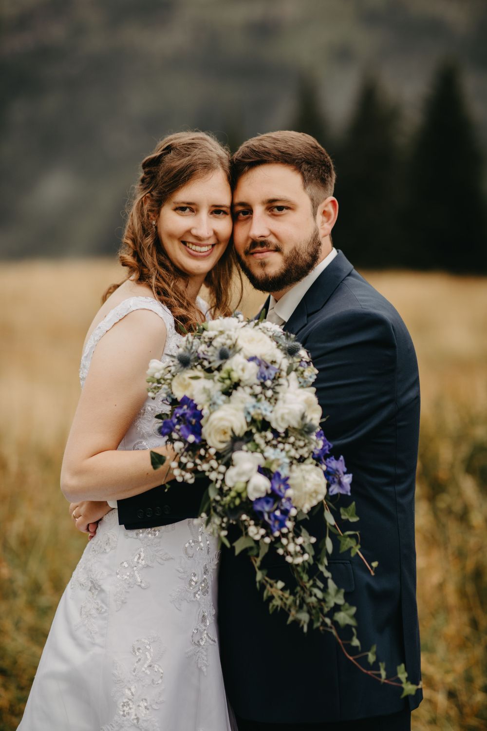Happy newlyweds pose with purple and white bouquet against mountain scenery.