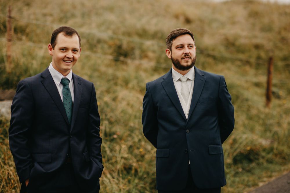Two groomsmen in suits smiling against mountain landscape.