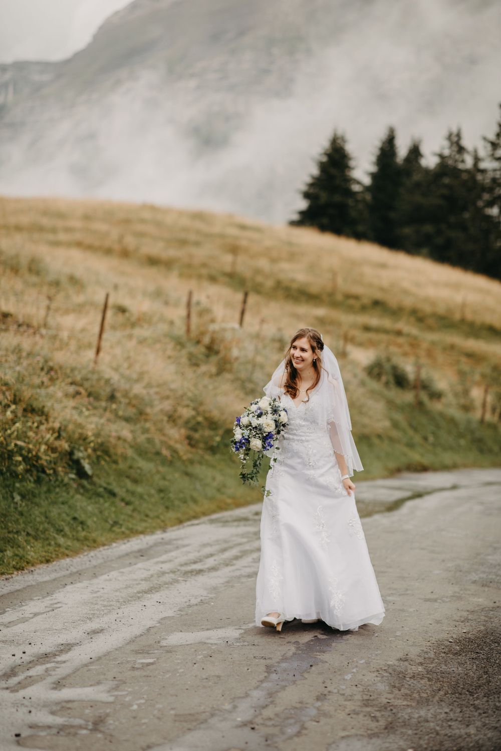 Bride in white gown holding bouquet on mountain road with misty peaks.