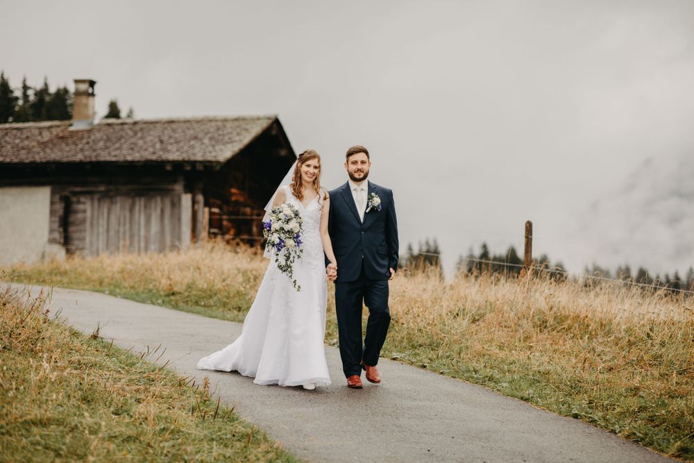 A couple walks along a mountain path on their wedding day with misty scenery and a rustic building in the background.