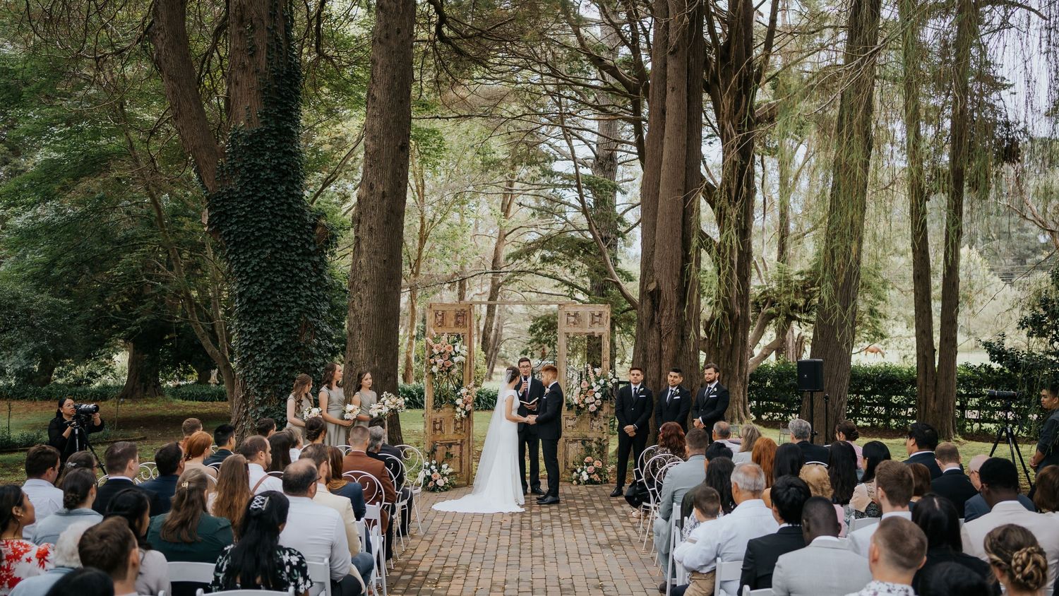 Outdoor wedding ceremony under towering trees with guests seated as couple stands at floral altar.
