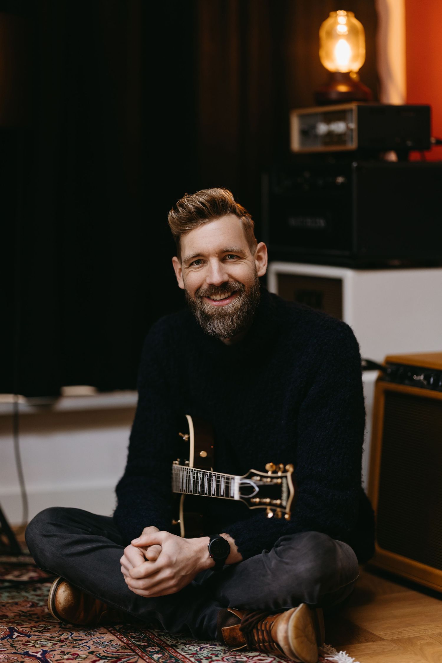Musician sitting cross-legged with acoustic guitar in cozy studio setting.