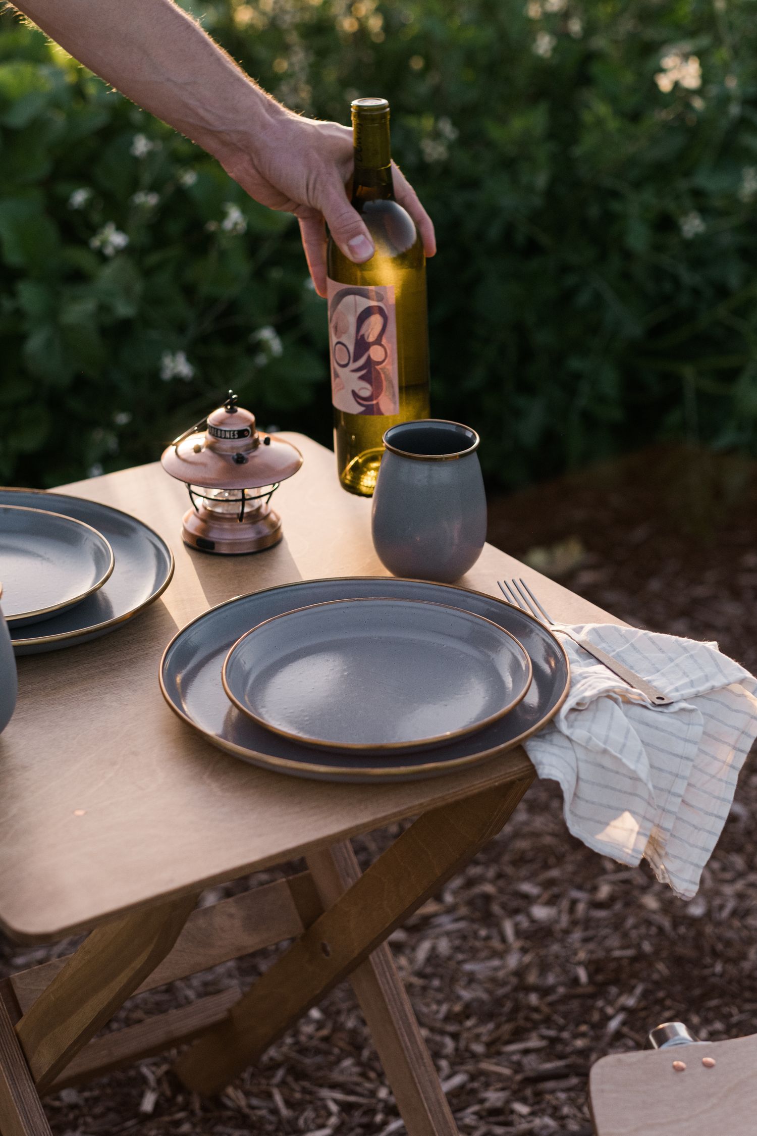 Hands setting up plates and wine on a wooden folding table in an outdoor garden setting.