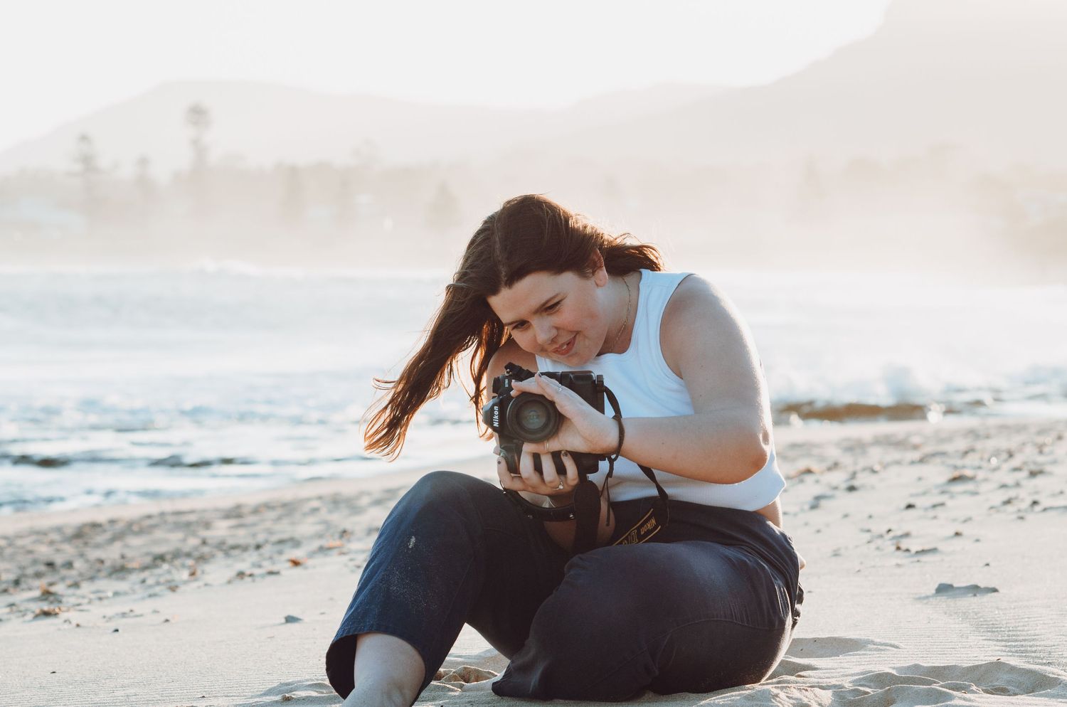 Photographer reviews images on camera while sitting on beach at sunset.