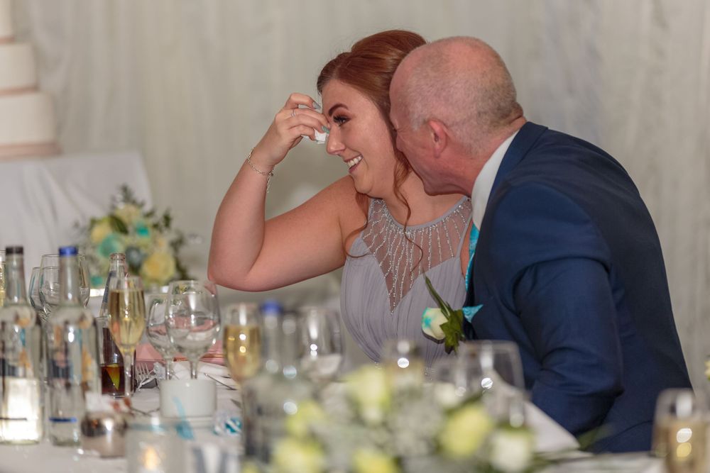 bridesmaid wipes a tear away from her eye comforted by the grooms father during the wedding speeches