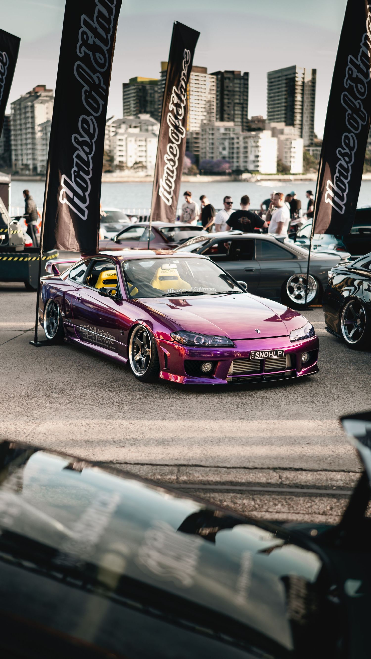 Modified purple Nissan Silvia sports car displayed at urban car show with city skyline backdrop.
